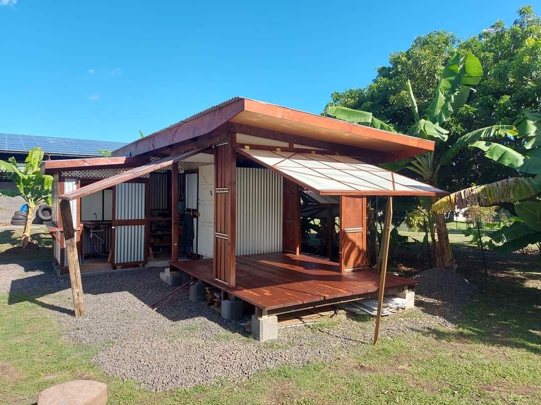 The bungalow features a wooden exterior with large overhanging eaves providing shade. A spacious deck extends outward, complemented by nearby greenery and banana trees. Solar panels are visible on the roof, highlighting a sustainable design in the natural landscape.