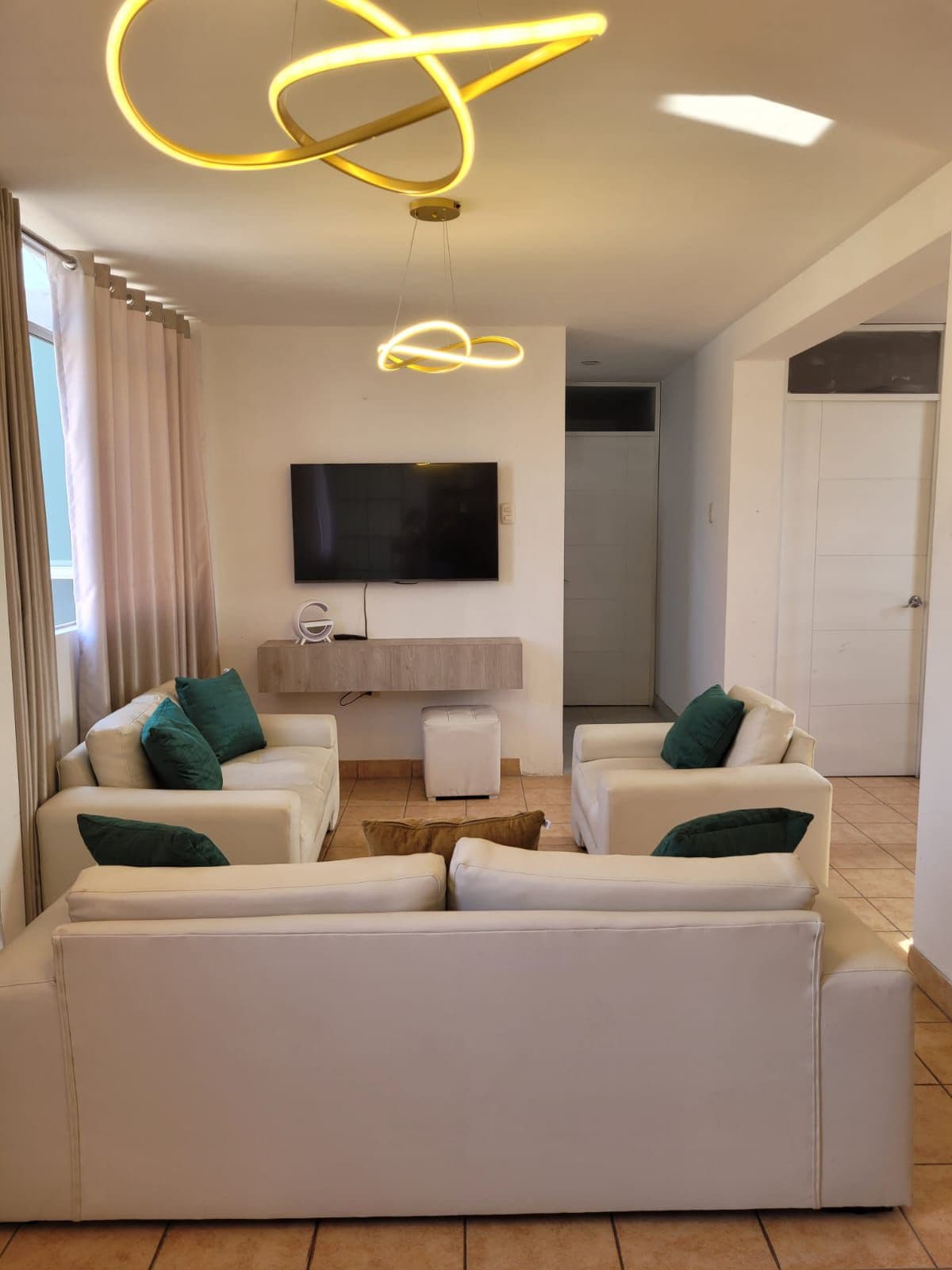 A living area is presented with four white armchairs arranged around a small wooden table. A wall-mounted television is visible above a light wooden console. Soft light is provided by a contemporary ceiling fixture. Natural light streams in through large windows covered by sheer curtains.