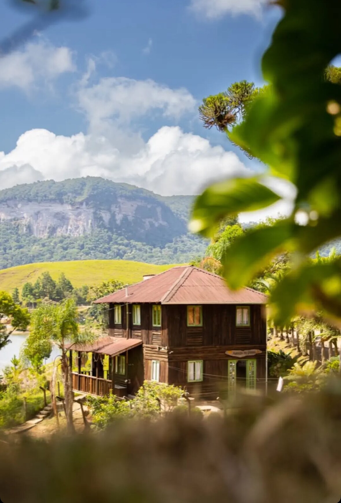 A charming wooden house is nestled amid lush greenery, with a backdrop of rolling mountains under a blue sky. Large windows and a covered porch overlook a picturesque landscape, providing a serene connection to nature.