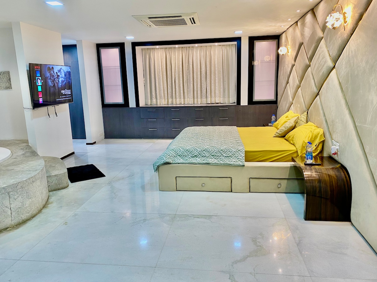 A spacious bedroom features a modern design with a light-colored bedspread and two yellow pillows. A wall-mounted television is positioned across from the bed, while a stylish grey accent wall adds texture. Natural light filters through a window with sheer curtains, enhancing the ambiance.
