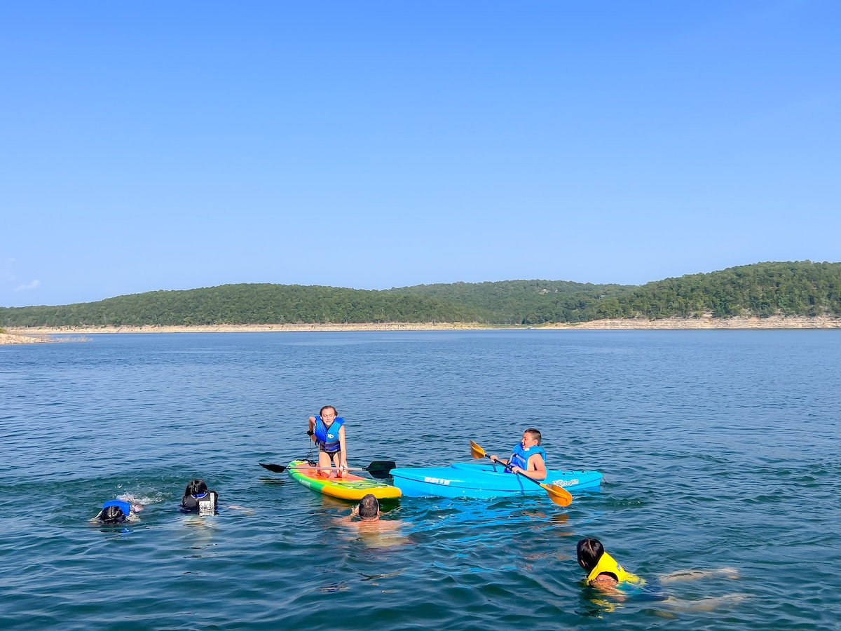 A bright day on the lake features individuals kayaking and swimming in clear water. Two kayakers are visible, one in a blue kayak and another in a yellow kayak, while swimmers in life jackets enjoy the water. Lush green hills serve as a scenic backdrop.