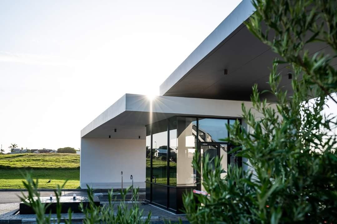 A modern architectural structure is showcased with sleek lines and expansive glass walls, allowing for natural light to flood the interior. Surrounding greenery frames the building, while the sun creates a soft glow on the roof, enhancing the contemporary design.