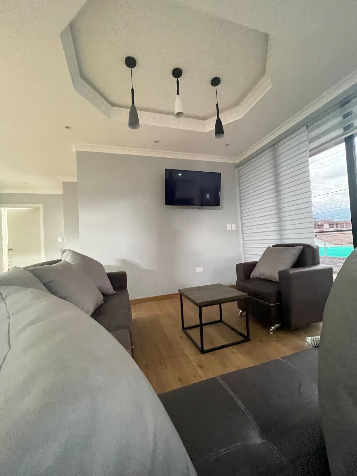 A modern living area is depicted, featuring a gray sofa and a dark leather armchair. A small black coffee table resides in the center, with a mounted flat-screen television above. Natural light filters through the tall windows adorned with soft blinds.