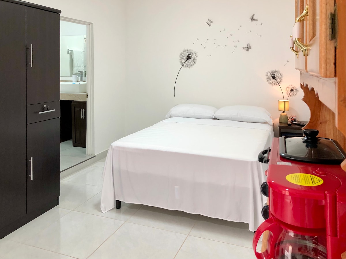 The sleeping area features a spacious bed dressed in a white coverlet. A simple bedside lamp provides soft lighting. A dark cabinet stands against one wall, while a red coffee maker is positioned on a surface nearby. A bathroom area is visible through an open door.