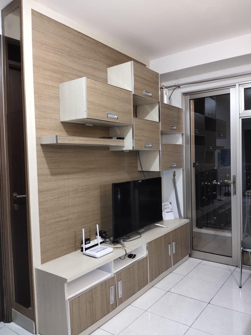 A modern living area is presented with a wall-mounted media unit featuring a television and various shelves. A sleek glass door provides access to a balcony, while light-colored cabinets offer ample storage. The overall design includes neutral tones that promote a calm environment.