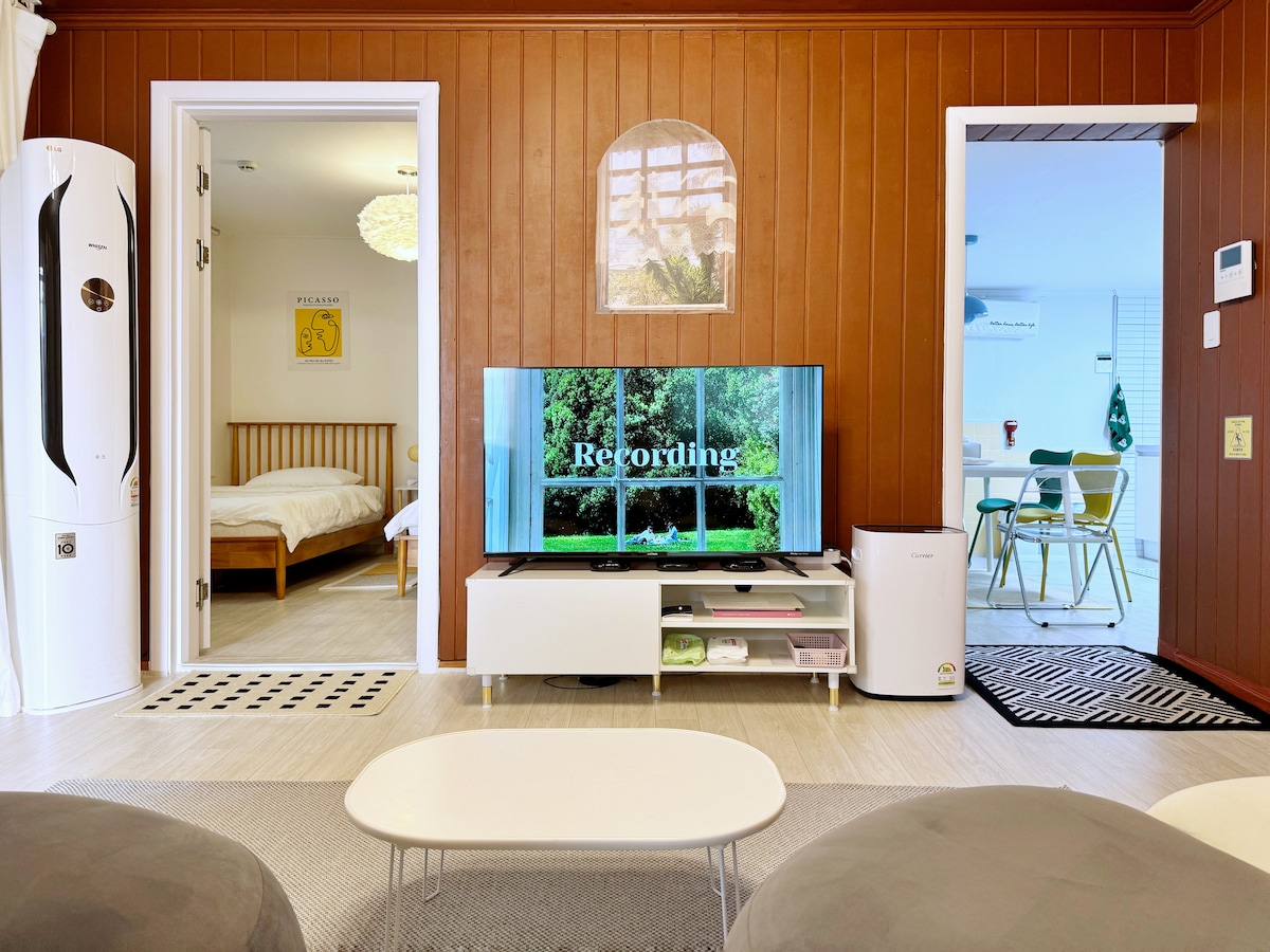 The living area is furnished with a low coffee table and a white entertainment unit displaying a screen. A cozy sofa and sleek air conditioning unit are positioned against warm wooden walls. Two doorways lead to additional rooms, creating a welcoming and open layout.