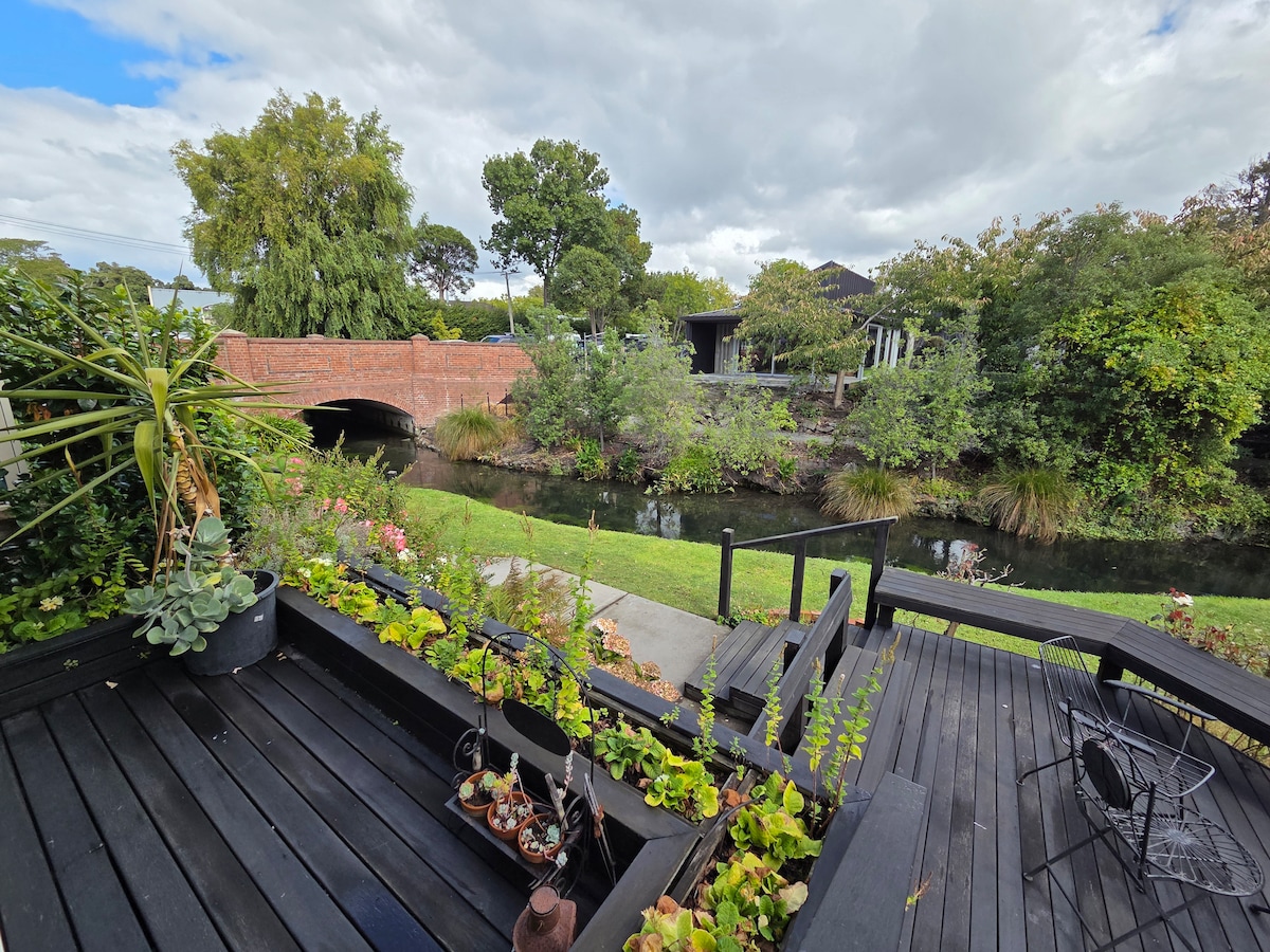 A wooden deck area is visible, adorned with various potted plants and greenery. A small, winding stream can be seen in the background, with a brick bridge crossing over it. Lush trees and shrubs create a serene, natural setting.