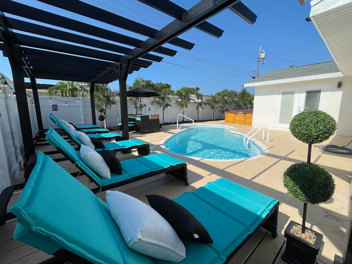A relaxing pool area is highlighted by a sparkling swimming pool surrounded by lounge chairs draped in vibrant turquoise fabric. A wooden pergola provides shade, complementing the sunny atmosphere. Lush greenery is visible, enhancing the serene outdoor space.