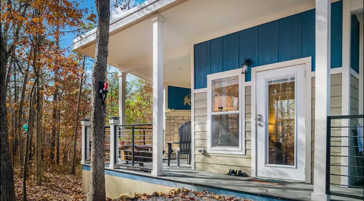 A quaint cottage exterior showcases a wraparound porch with seating, framed by tall trees and autumn foliage. The entrance is highlighted by a glass door, inviting guests to explore the cozy interior. Soft natural light filters through the space, enhancing the peaceful surroundings.