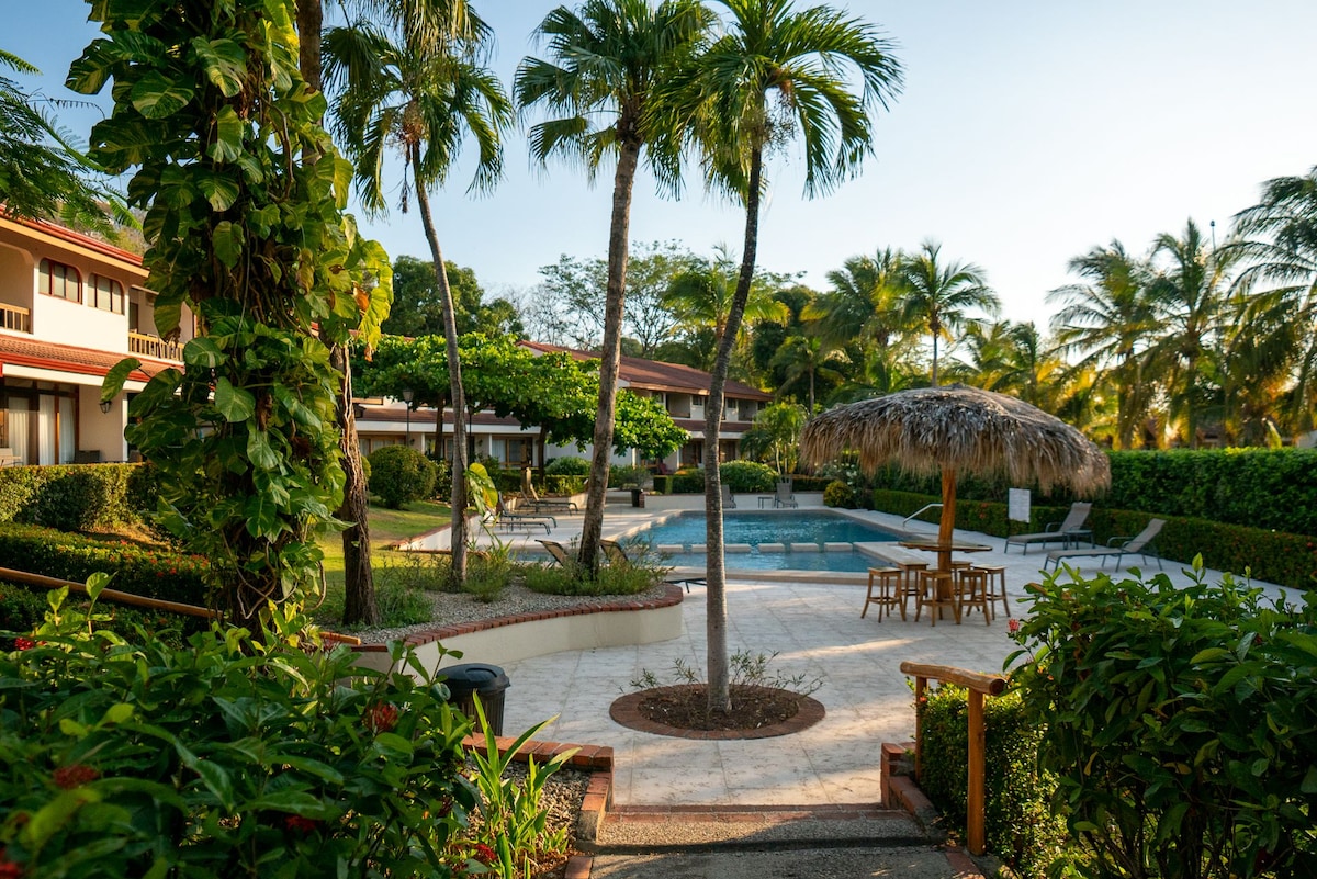 The serene outdoor area features a swimming pool surrounded by palm trees and well-maintained gardens. A thatched-roof umbrella provides shade over a seating area with wooden chairs, inviting relaxation in the tranquil environment.