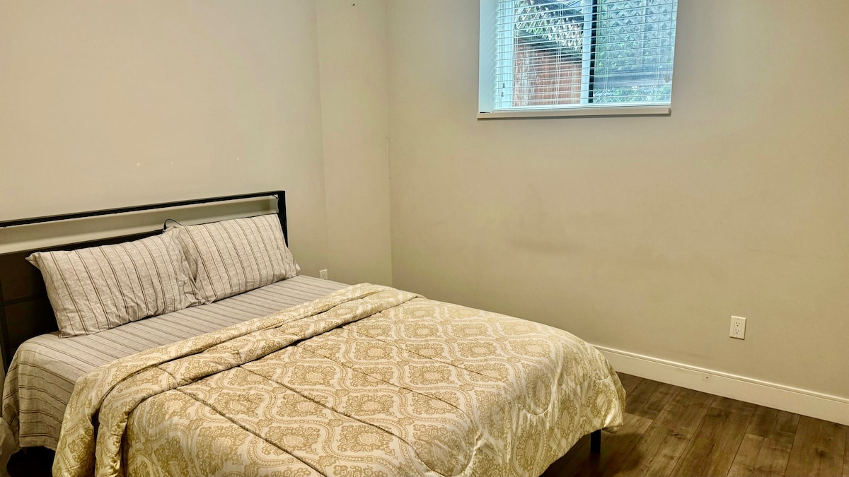 A private room features a spacious bed covered with a patterned blanket. Light enters through a window, illuminating the neutral-toned walls and floor. The minimalist decor enhances the calm ambiance of the space.