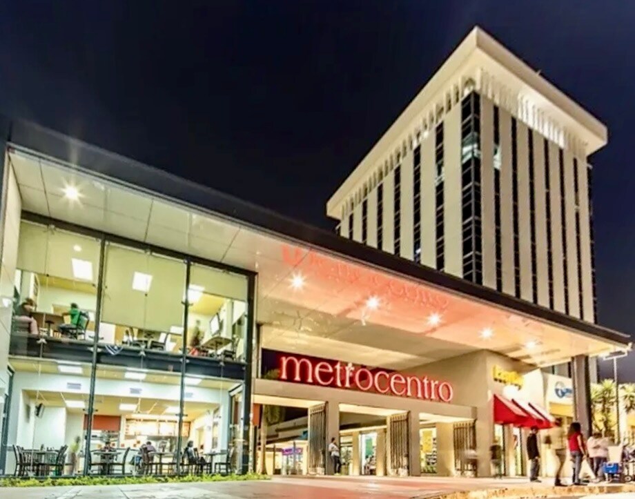 The exterior of the Metrocentro shopping center is depicted, showcasing a modern design with large glass panels. Bright signage displays the name 'Metrocentro,' and various shops are visible through the entrance. The area is well-lit, creating a welcoming atmosphere during the evening.