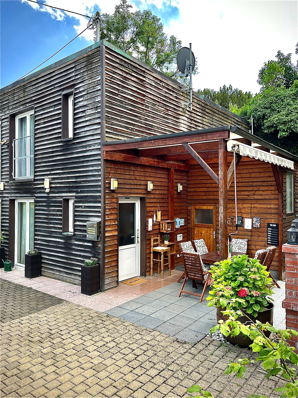 The exterior of a modern wooden house features a covered seating area with a wooden table and chairs. The entrance door is framed by a small patio, while greenery surrounds the building. Sky and trees can be seen in the background.