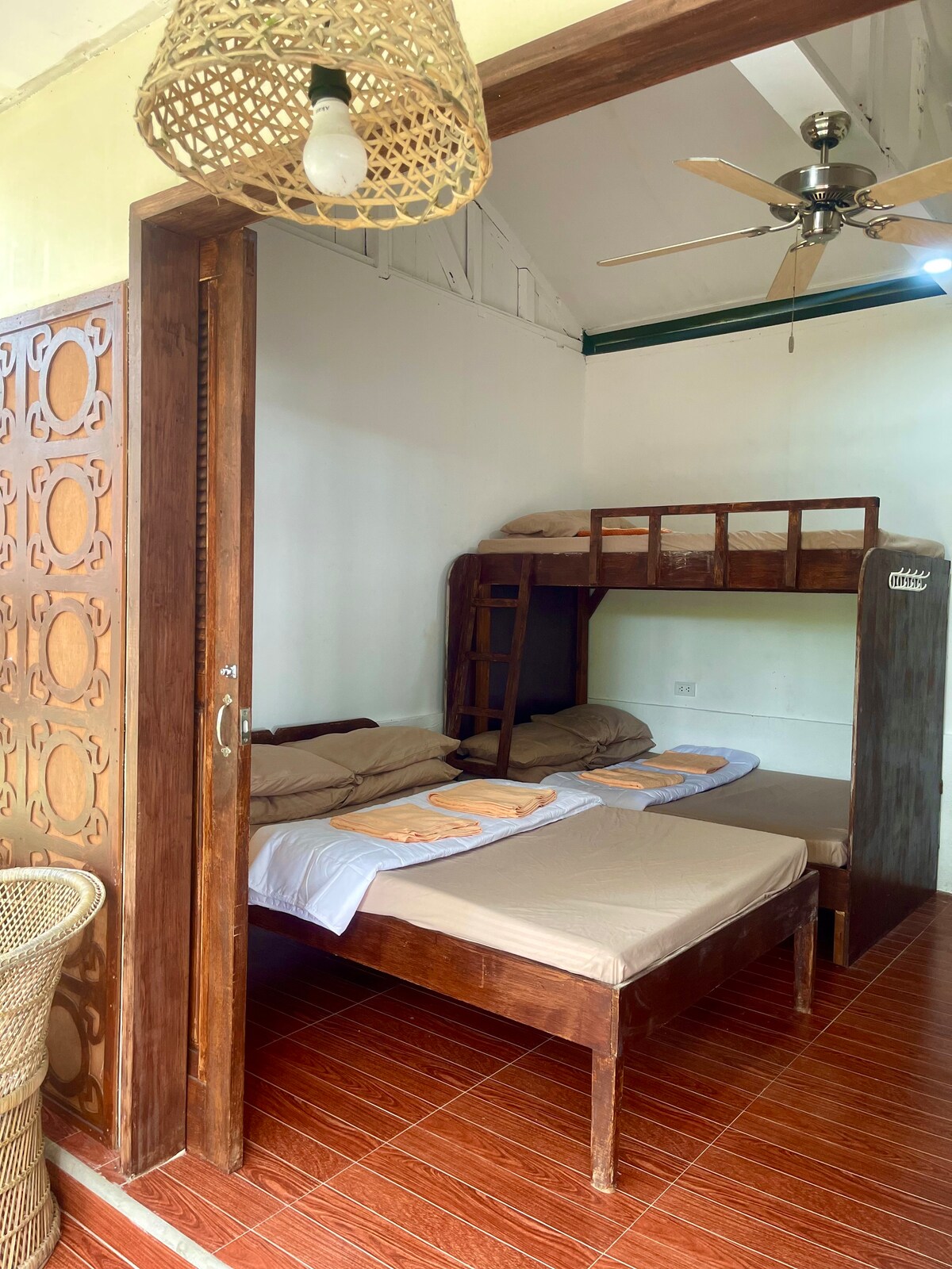 A cozy room features a combination of single and bunk beds, with beige bed linens neatly arranged. The room is illuminated by natural light coming through the open doorway. A ceiling fan is visible above, enhancing the relaxed atmosphere.