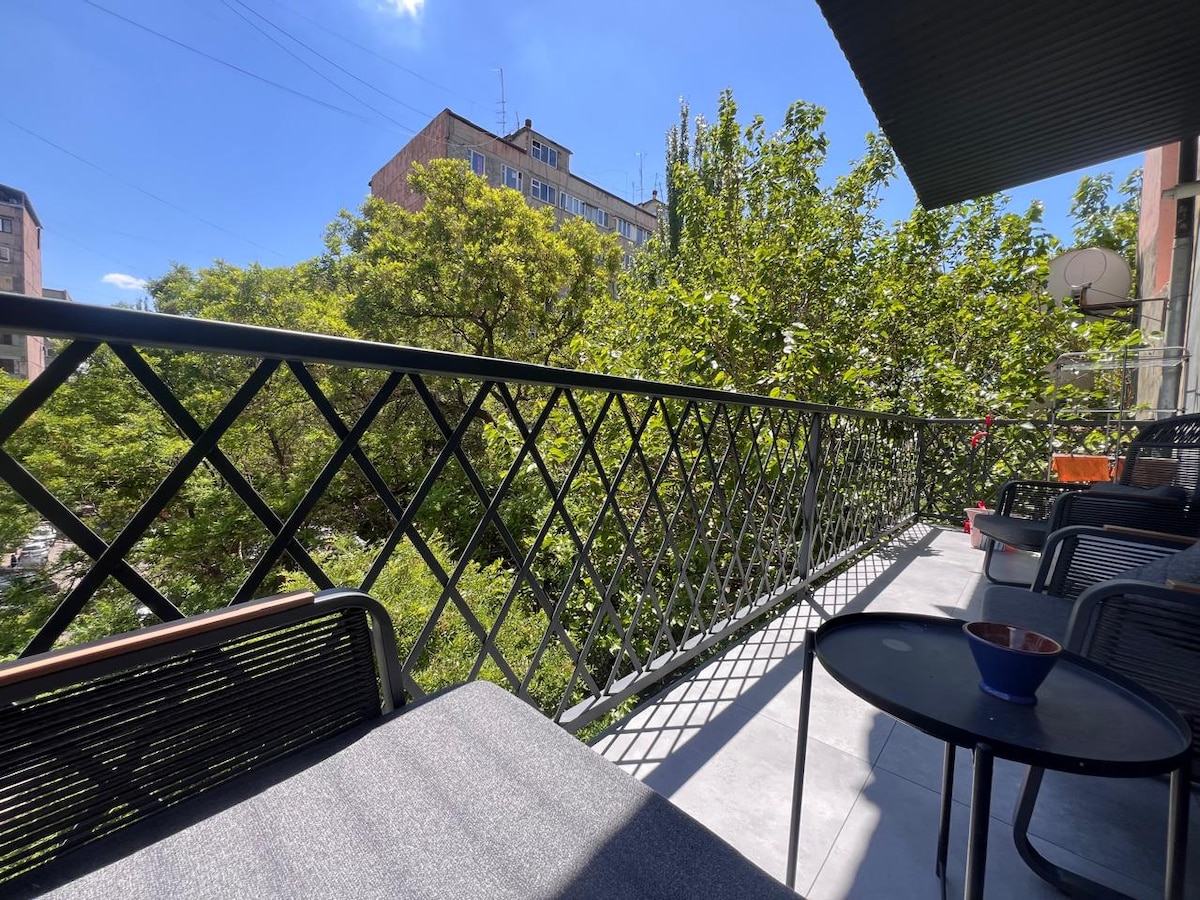 A balcony is presented, offering views of lush green trees and neighboring buildings under a clear blue sky. Black metal furniture, including chairs and small tables, is arranged thoughtfully, creating a welcoming outdoor space for relaxation.