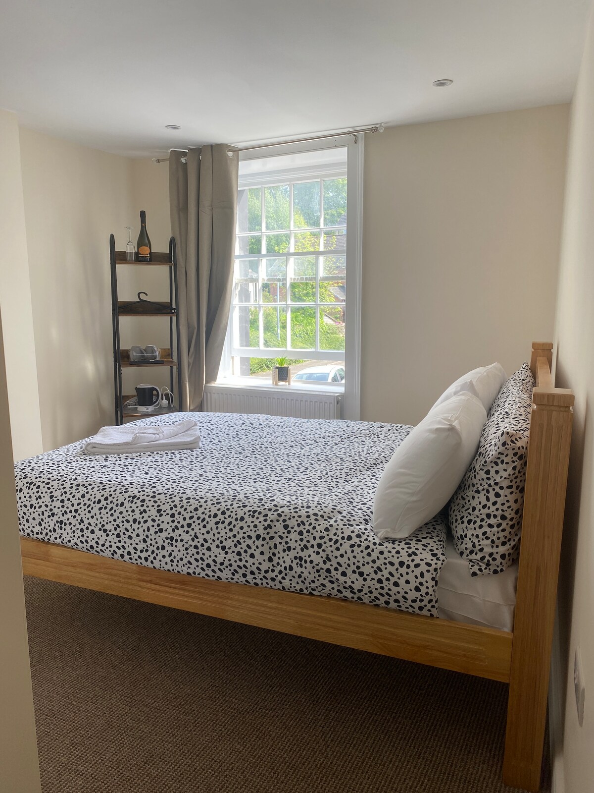 A bright bedroom features a spacious bed dressed in a patterned white and black duvet. A window introduces natural light, and a small shelving unit presents beverages and cups. Neutral walls and carpeting enhance the room's simplicity and warmth.