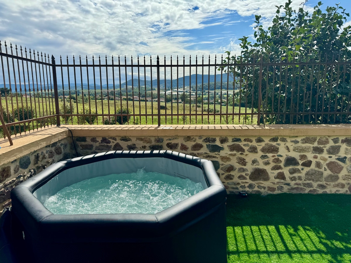 A well-maintained jacuzzi is set against a backdrop of rolling hills and a clear sky. The fenced area, featuring stone walls, provides privacy while offering a view of the surrounding landscape, complemented by patches of green grass.