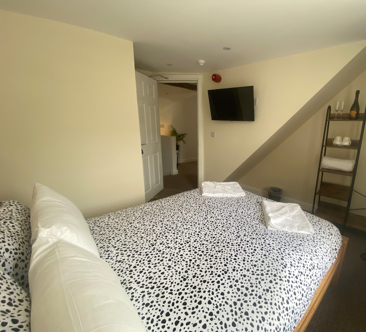 A spacious bedroom features a large bed adorned with a spotted bedspread. Fresh towels are neatly arranged on the bed. A flat-screen television is mounted on the wall, and a staircase is visible in the background. The room is well-lit with soft, neutral walls.