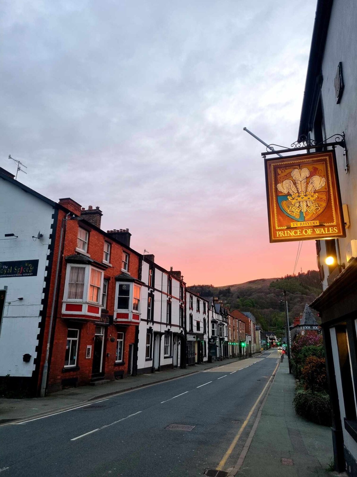 The image depicts a quiet street scene in Llangollen, with traditional buildings lining both sides. A pub sign reading 'Prince of Wales' is visible on the right, while the natural landscape is seen in the background under a softly lit sky at dusk.