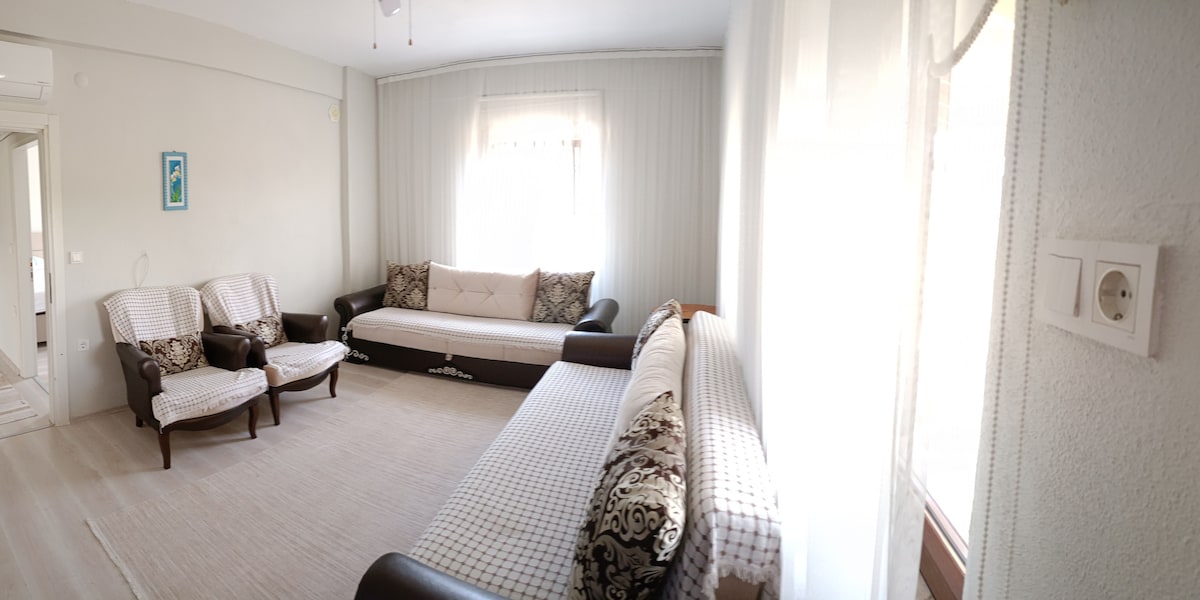 A spacious living area features light-colored walls and a soft, neutral carpet. Seating includes two patterned armchairs and a large, convertible sofa with decorative cushions. Large windows allow natural light to fill the room, creating a bright ambiance.