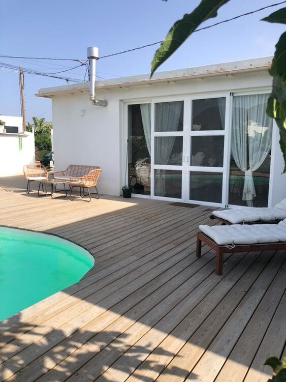 A wooden deck surrounds a pool with a soft green hue, leading to a house featuring large glass doors and light curtains. Two woven chairs are positioned on the deck, complemented by a lounge chair, creating an inviting outdoor seating arrangement.