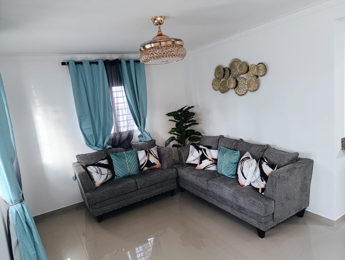 A bright living room features two gray sofas adorned with patterned cushions in turquoise and black. Decorative wall art made of woven materials adds texture to the space. Natural light filters in through sheer curtains, complementing the overall serene ambiance.