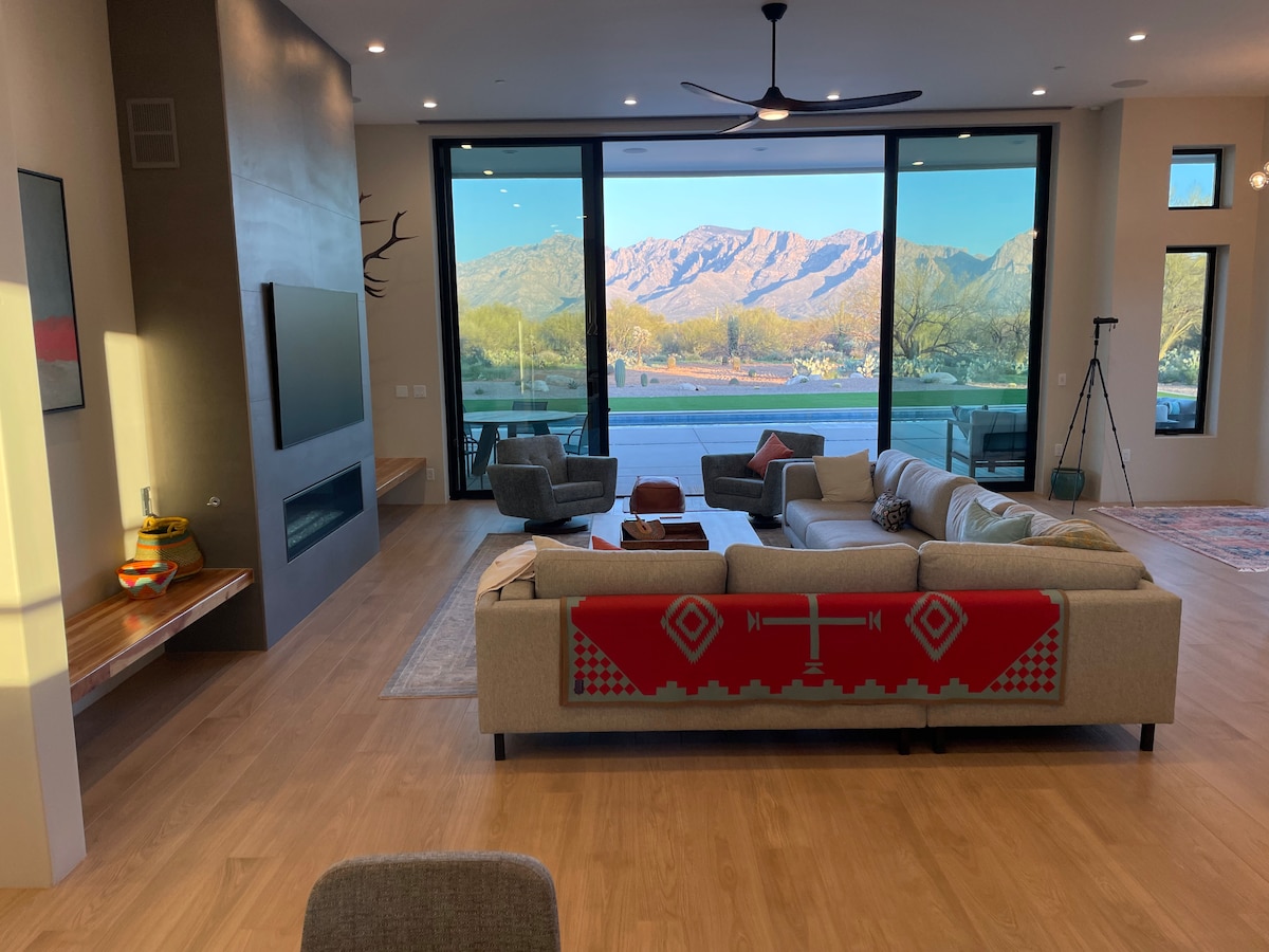 An open living area showcases large glass windows framing stunning views of the surrounding mountains. A comfortable light-toned sofa is draped with a geometric throw, complemented by modern armchairs and a minimalist coffee table. Natural light fills the space, enhancing the overall ambiance.