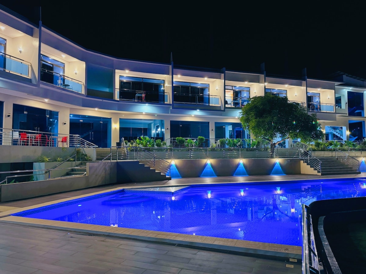 A modern retreat is illuminated at night, showcasing a striking blue pool that reflects the surrounding light. The well-maintained outdoor area features a combination of greenery and paved surfaces, while multiple balconies offer views of the pool and the landscape.