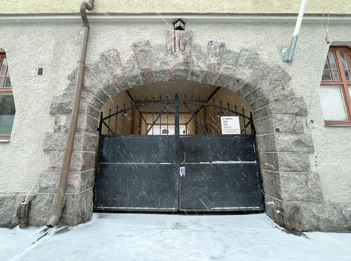A sturdy stone archway frames a large black gate, providing access to the building's interior. The gate is detailed with elegant ironwork and is set against a textured, pale grey wall. Snow is visible on the ground, creating a serene winter scene.