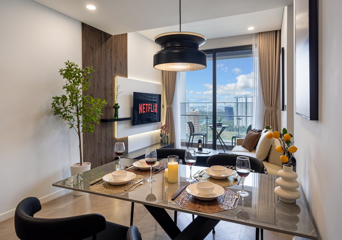 A dining area is presented with a sleek glass table set for four, showcasing dishes, cutlery, and wine glasses. A bowl of decorative items adds a touch of elegance. A modern TV mounted on the wall offers access to streaming services, while large windows reveal outdoor seating and a city view.