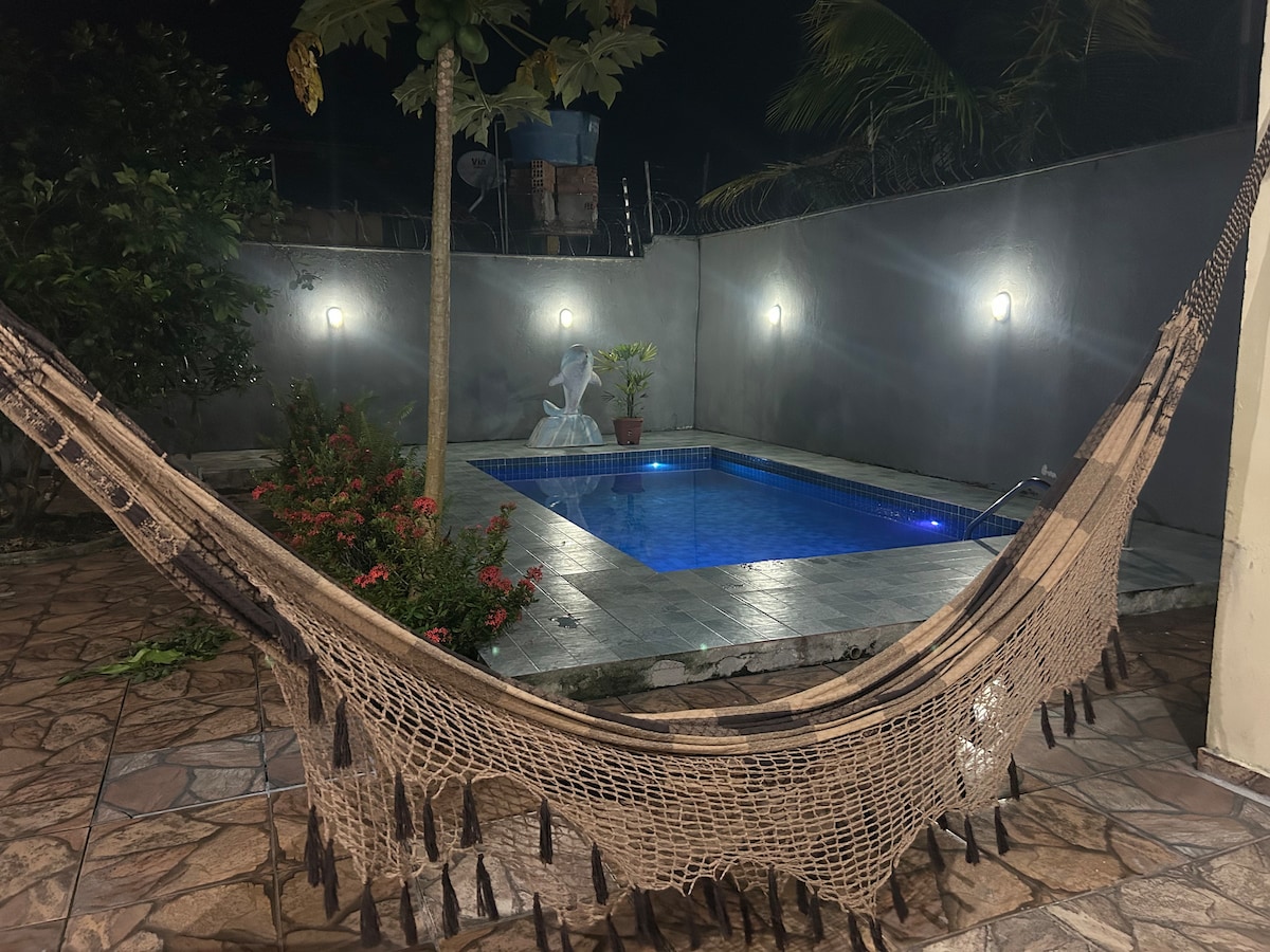 A serene outdoor space is showcased at night, featuring a tranquil blue pool surrounded by soft garden lighting. A cozy hammock is positioned in the foreground, adding a relaxed vibe to the area with tropical plants and a mural-styled wall visible in the background.