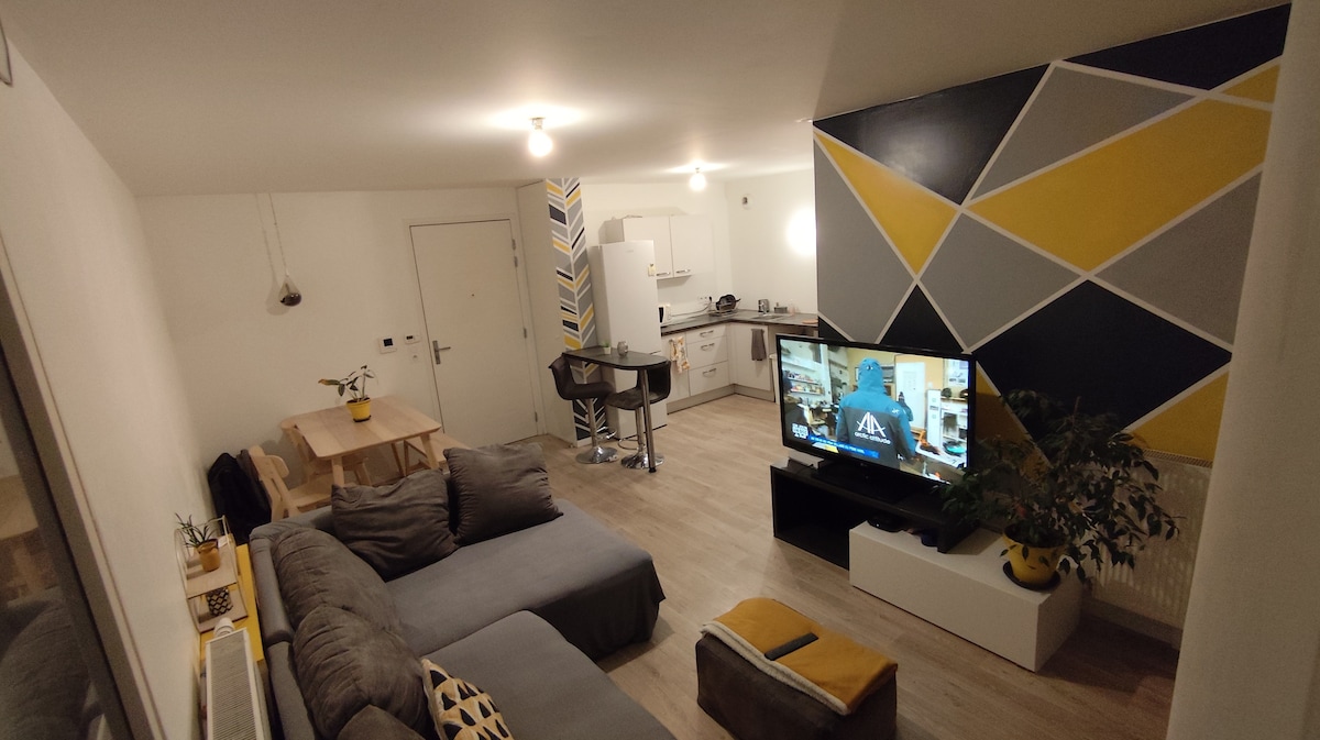 A spacious living area features a contemporary sectional sofa arranged around a low coffee table. A large television sits on a white media unit. Open kitchen space is visible in the background, with modern appliances and vibrant geometric wall accents adding a touch of color.