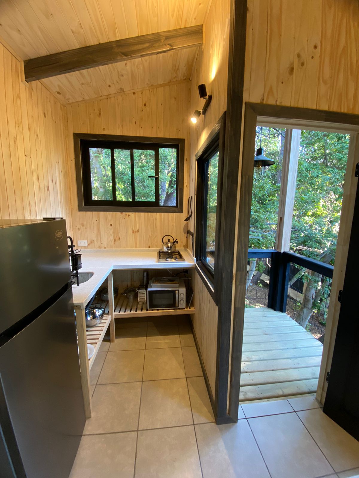 An inviting kitchen area features wooden walls and a tiled floor, providing a warm ambiance. Equipped with modern appliances, including a refrigerator and a stovetop, it offers views of the surrounding greenery through large windows. A balcony extends outwards, enhancing the connection with nature.