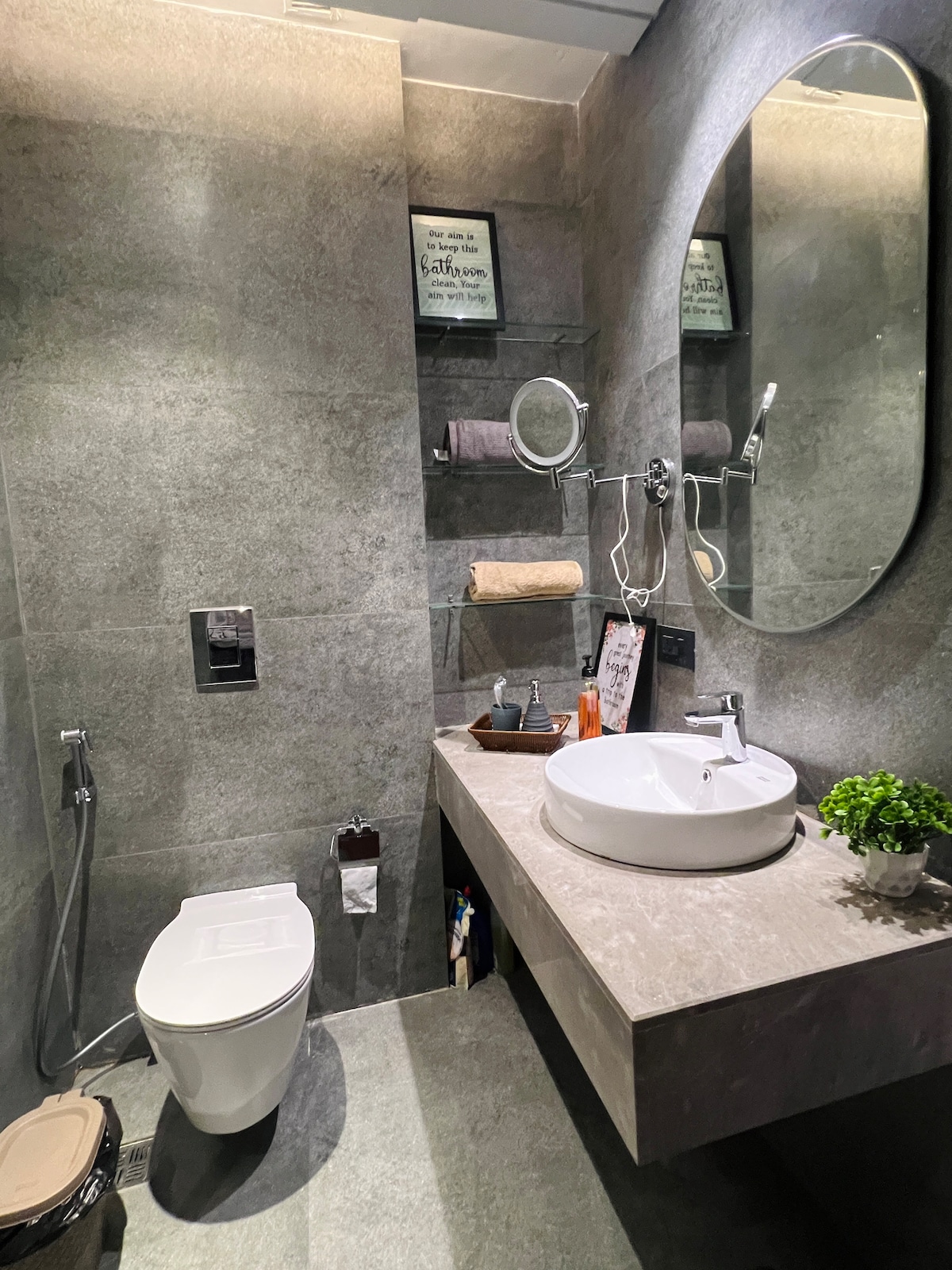 A modern bathroom features sleek gray tiles and a round mirror above a minimalist sink. A contemporary toilet is positioned nearby, with a towel rack displaying neatly arranged towels. Decorative items are placed on the sink, adding a touch of style to the functional space.