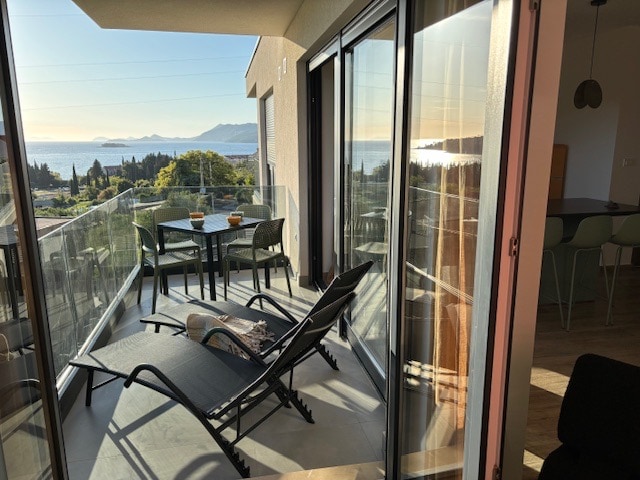 A spacious balcony offers a sea view, featuring two lounge chairs and a small dining table set for four. Large glass doors provide an unobstructed view of the surroundings, while natural light fills the area, enhancing the connection between indoor and outdoor spaces.