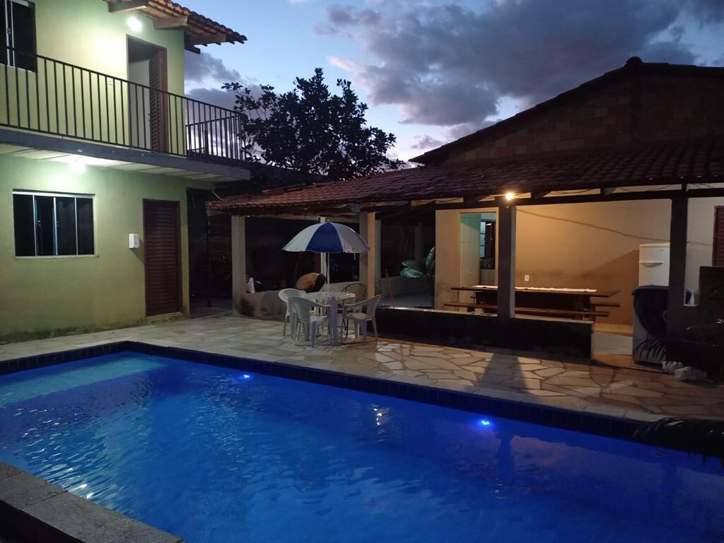 A tranquil outdoor area features a solar-heated swimming pool with illuminated blue lighting. A shaded seating area is accommodated by a patio umbrella, with a dining table nearby. Surrounding spaces include a two-story house, highlighting a harmonious blend of indoor and outdoor living.