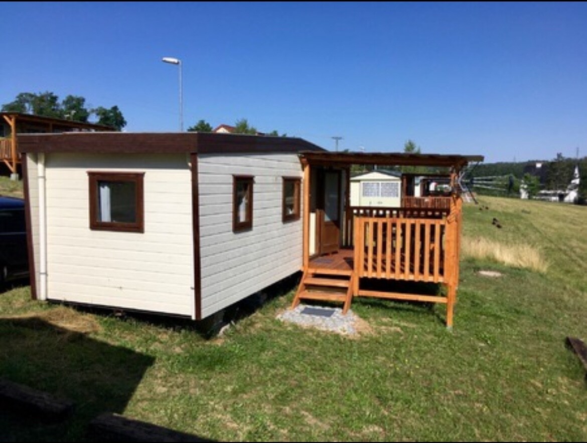 A small cabin is shown with crisp white siding and wooden accents. A wooden deck featuring steps leads to the entrance of the cabin. Surrounding greenery is visible, highlighting the serene outdoor environment.