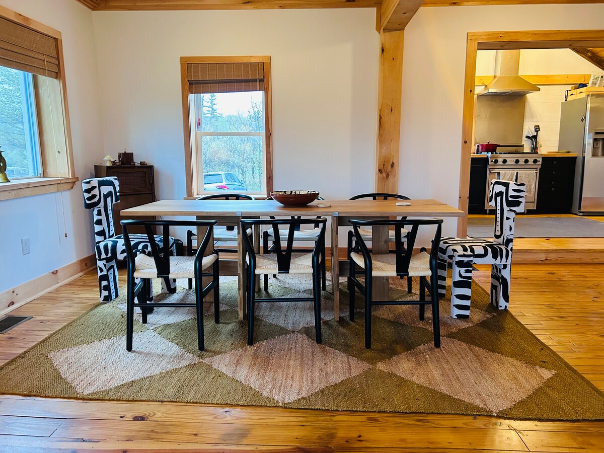 A spacious dining area showcases a long wooden table surrounded by black and white patterned chairs. Natural light fills the room through the large windows, illuminating the warm wood floors and unique area rug beneath the table.