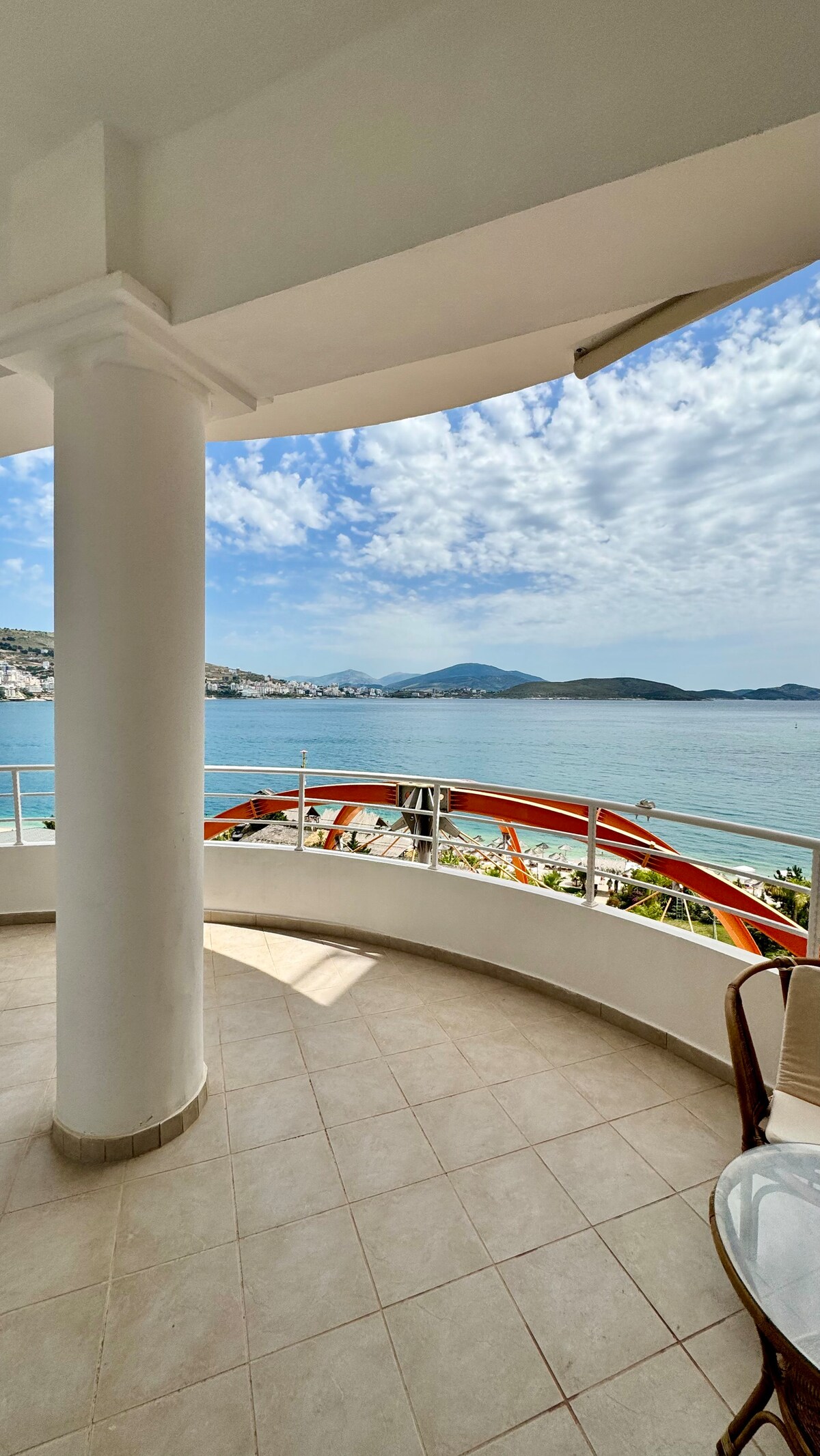 A private balcony offers unobstructed views of the Ionian Sea, framed by a circular design. Coastal hills are visible in the distance, while a bright sky with scattered clouds enhances the serene atmosphere. A small table and chairs provide a comfortable spot for relaxation.