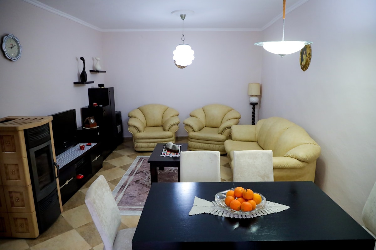 A cozy living area is furnished with two light-colored sofas and a dark wood coffee table. A flat-screen TV is positioned on a black storage unit. A dining table with white chairs displays a bowl of oranges, adding a touch of color to the space.
