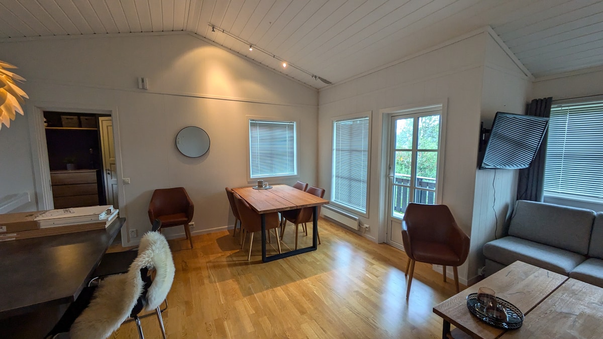 A bright living and dining area features large windows allowing natural light to fill the space. A wooden dining table is surrounded by six chairs. A cozy seating arrangement includes a grey sofa and two armchairs. Soft lighting and natural wood accents enhance the ambiance.