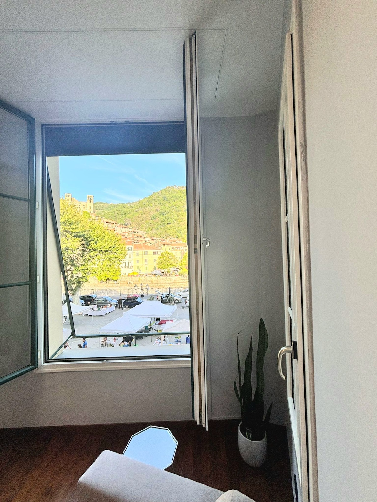 A bright interior features an open window that invites fresh air and light. Outside, a view of lush green hills and a bustling market square can be seen. A tall plant in a simple pot adds a touch of nature to the space.