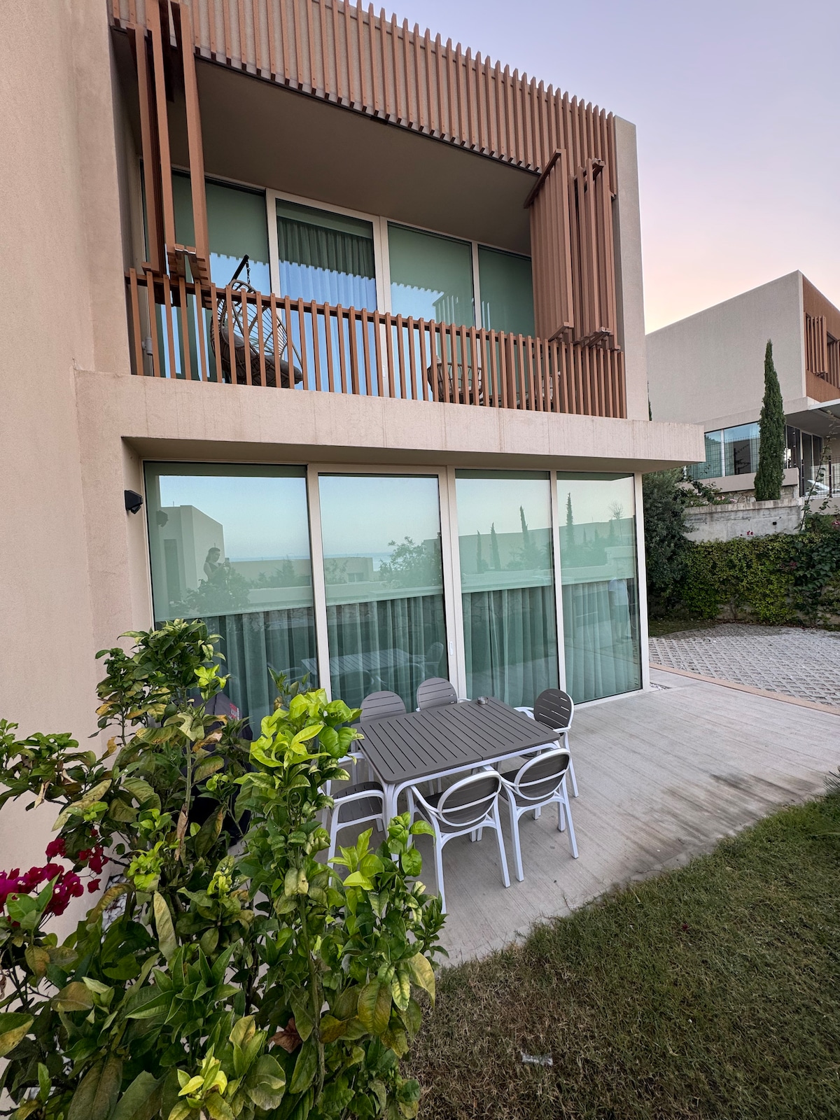 An outdoor dining area is situated on the terrace, featuring a gray table surrounded by several white chairs. Large glass doors offer a view into the villa, while lush greenery and flowering plants frame the scene, enhancing the connection with nature.
