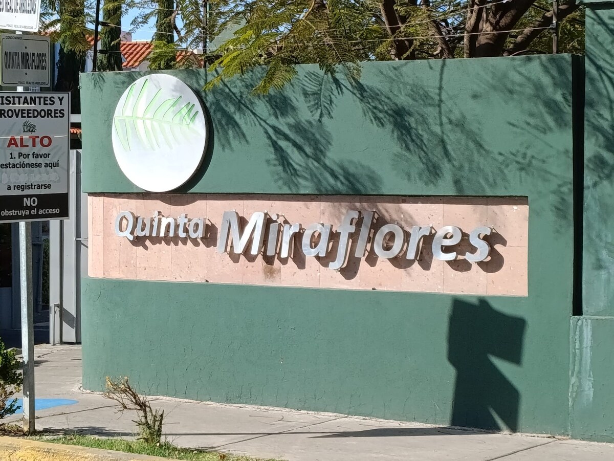 A green wall features a prominent sign with the name 'Quinta Miraflores' displayed in modern lettering. The natural ambiance is enhanced by surrounding trees, creating a welcoming entrance to the property.
