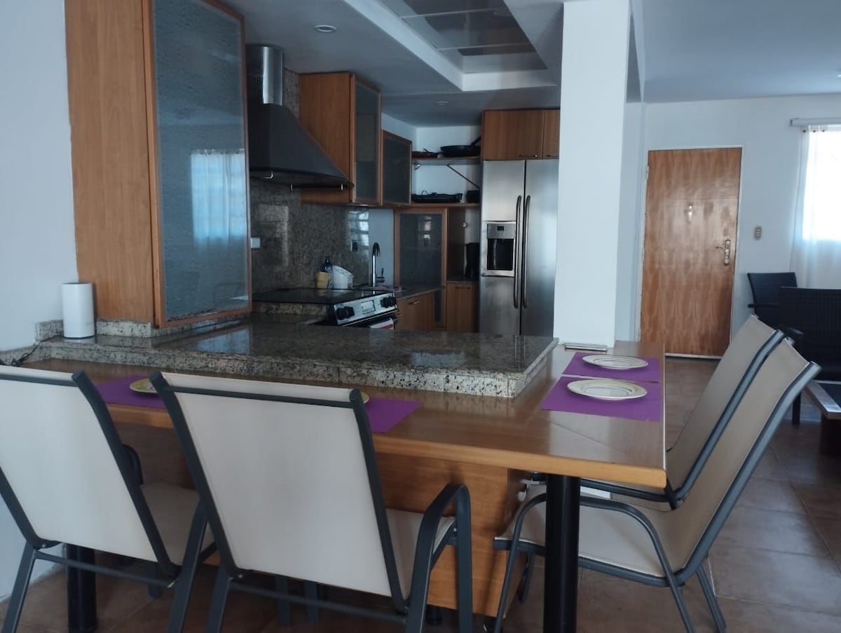 A modern kitchen area features wooden cabinetry and granite countertops. A spacious dining table, surrounded by four metal-framed chairs, is set with purple placemats. Stainless steel appliances, including a refrigerator and an oven, are visible along with a sleek range hood.