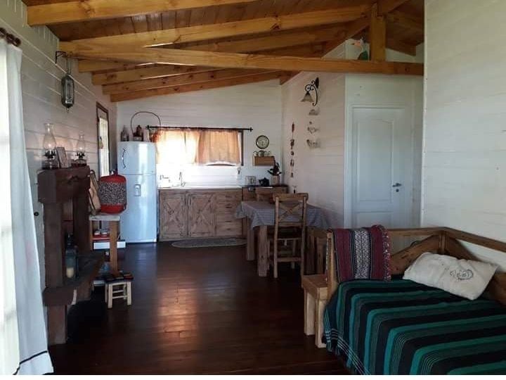 An open living area features wooden beams and a warm, inviting color palette. A kitchen is visible with a white refrigerator, complemented by a dining table and wooden chairs. A cozy seating arrangement is arranged with a striped blanket draped over a bench.