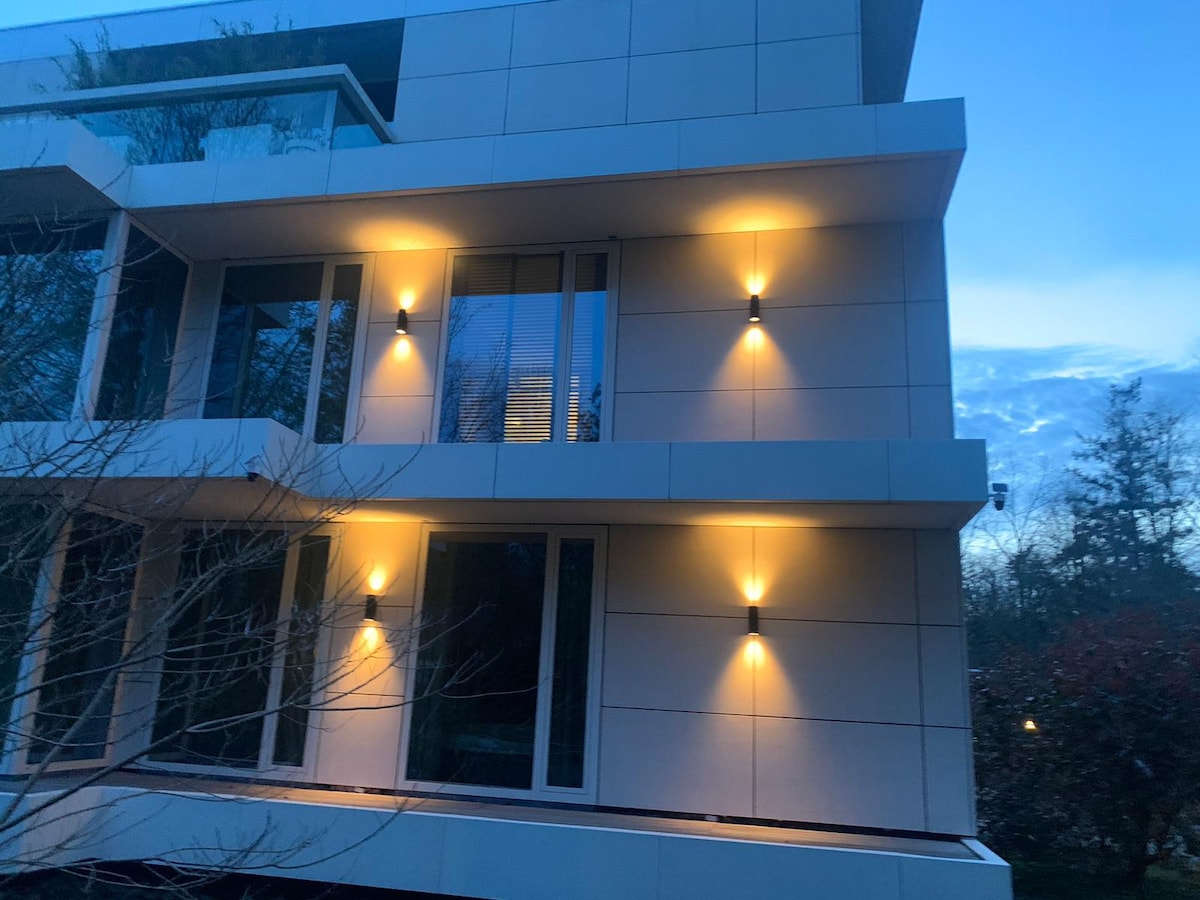 The exterior of the modern building is illuminated by warm wall lights, highlighting the sleek design and large glass windows. The evening sky provides a soft backdrop, creating a tranquil atmosphere around the property.