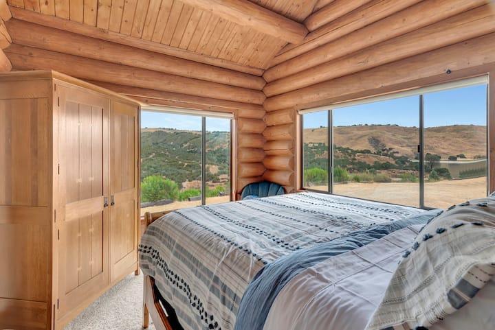 Bright and airy bedroom with dual hillside views—comfortable king bed, authentic log cabin construction with vaulted timber ceilings, ample closet space, and peaceful natural surroundings