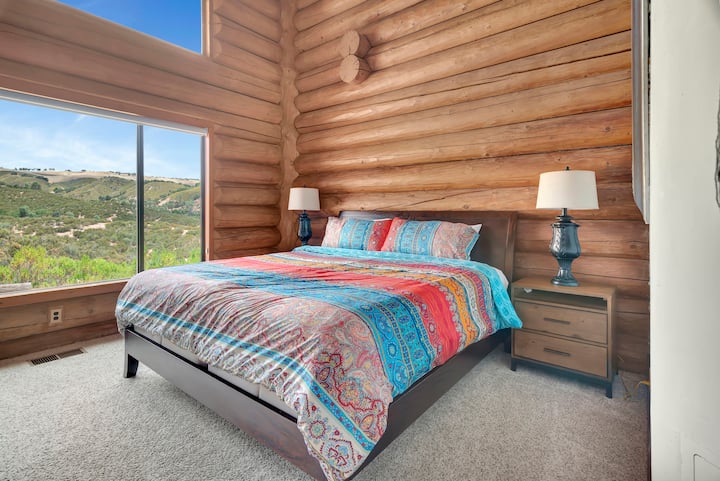 Ensuite bedroom with stunning lake views—king bed with vibrant linens, vaulted timber ceilings, and panoramic windows overlooking Lake Nacimiento