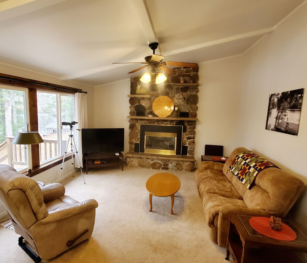 A cozy living area features a fireplace framed by natural stone, creating a warm focal point. Comfortable seating includes a plush sofa and armchair, complemented by a round coffee table. Large windows offer views to the outside, with natural light enhancing the neutral decor.